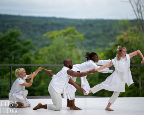 Four David Dorfman Dance dance in a group against the lush greenery of Jacob’s Pillow Henry Lier Stage. Nik Owens delicately holds the foot of Claudia Lynn-Rightmire as Rightmire eases into a supported lean. Kashia Kancey is just behind the duet, extending her right leg behind her as she gestures both arms forward. Nik is a man with a darker complexion and a shaved head and Claudia is a woman with blonde hair in an updo. Kashia is a woman with black hair tied in a ponytail and has a darker complexion. To the left of the trio crouches Lily Gelfand, a woman with short blonde hair, with arms softly bent before her, as if to reach towards the activity in front of her. All dancers don flowy white costumes.