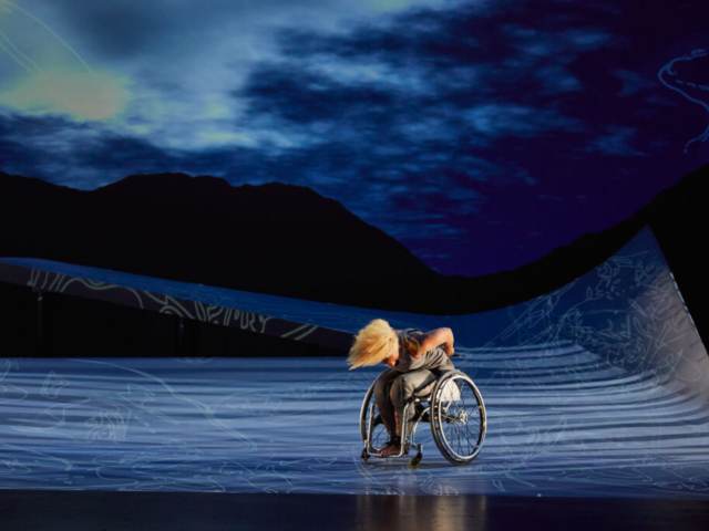 Alice Sheppard spins in her wheelchair, folded at the waist, arms behind her, blonde hair flying. She is a multiracial Black woman with coffee colored skin; she wears a gray bodysuit. A huge ramp surrounds her, Rodin-inspired figures are projected across the cold gray stage and a cloudy blue sky appears behind her. Photo Jaqlin Medlock / Rutgers University.