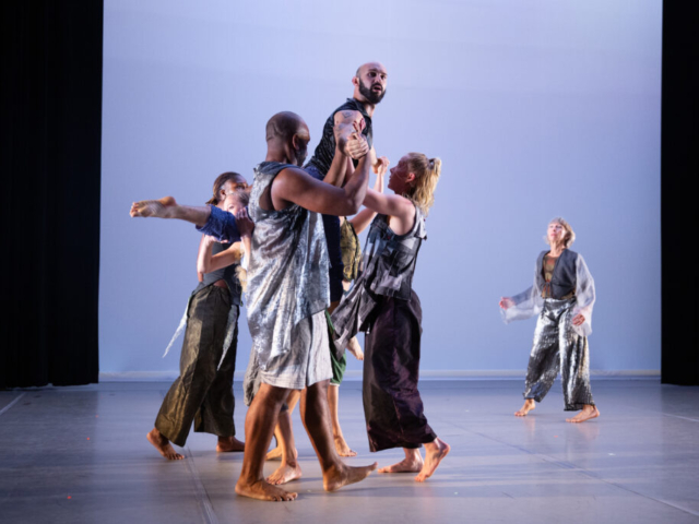 David Dorfman Dance dancers lift Jack Blackmon, a dancer with a brown-black beard clad in crocodile print pants and a leather vest. They gaze out at the audience, perched atop the company dancers on a brightly lit stage.