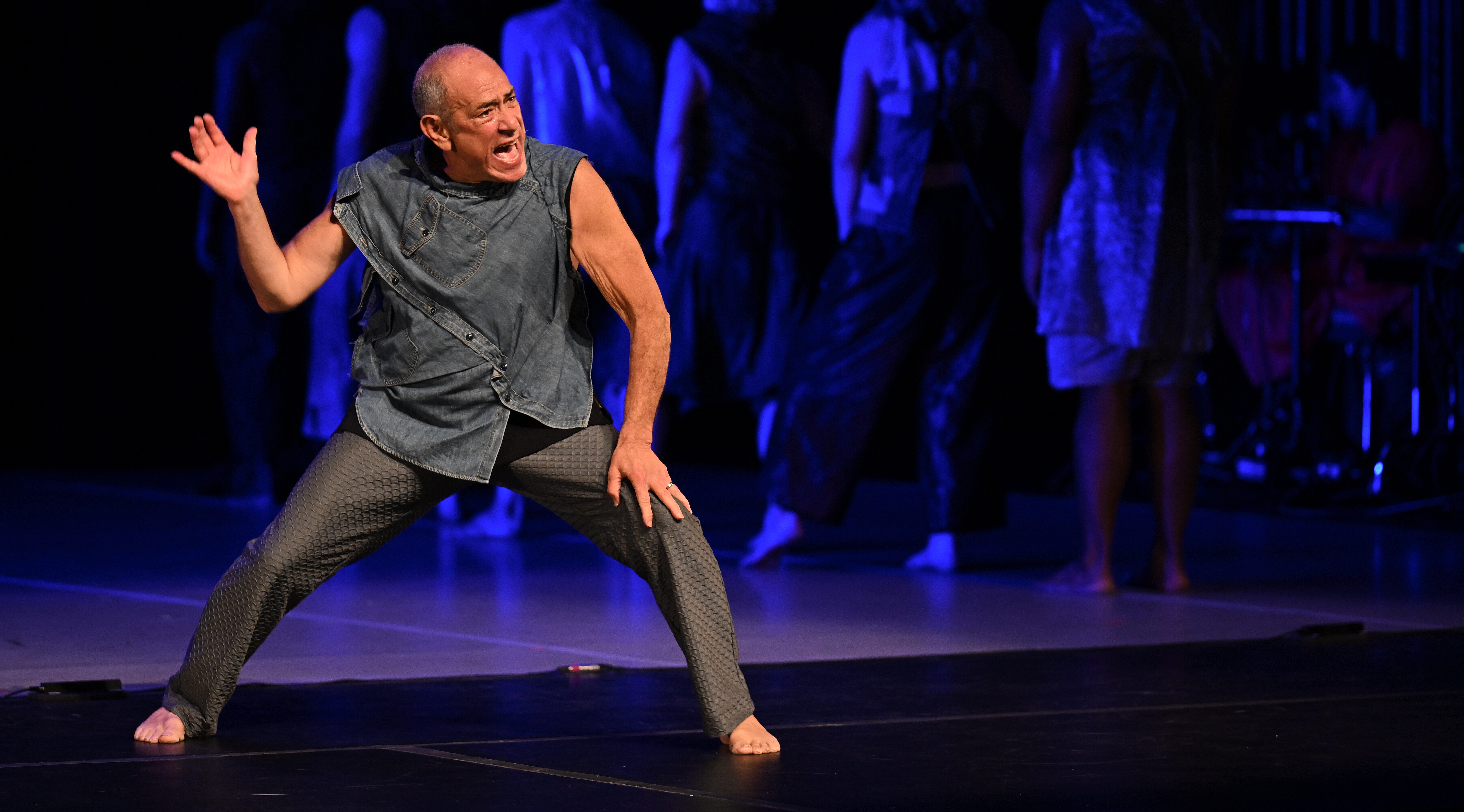 David Dorfman yells on stage, rearing back his hand in a shallow lunge, as if about to strike. Clad in an armor of denim tunic and silver scaled pants, he glares into the audience as a line of dancers are blurred in the purple background. David is a fair complexioned man with a shaved head.