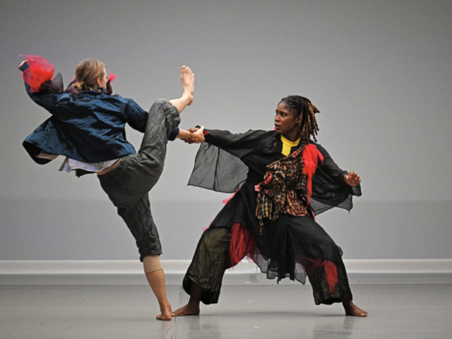 Kashia Kancey counterbalances Michaela Ellingson in a daring duet, as Michaela gestures her leg over their clasped hands. Weighted and grounded, Kashia looks to Michaela as Michaela faces away from the camera. Kashia is a dark complexioned woman with brown hair in an updo; Michaela is a woman with a fair complexion with dark blonde hair in a bun. Both don apocalyptic garb - mixed textures of wrinkled black, red flannel, and navy shirts and pants.