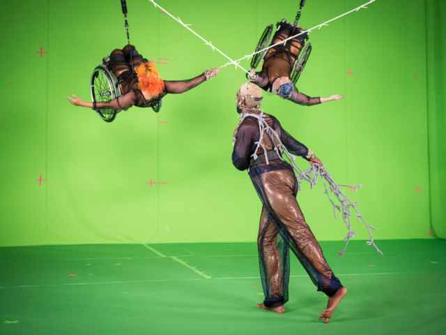 Alice Sheppard and Laurel Lawson soar in their wheelchairs in front of a bright green screen, each facing the ceiling, arms curved up, holding a white barbed wire prop in the shape of a huge X.  Jerron Herman faces the duo, his back to the camera, one leg stretched behind him and the opposite arm out to the side holding a cloth wire prop whip which wraps his body.  Alice is a multiracial Black woman with short bright orange curly hair and coffee-colored skin; Laurel is a white person with pale skin and cropped peacock blue hair; Jerron is a Black man with a dark beard and blonde shortcut hair. They all wear copper bodysuits overlaid with black mesh which shimmers in crimson, sapphire, and amethyst, and delicate filigree headpieces of copper and brass with pearl and gold beading. Photo by Cherylynn Tsushima.