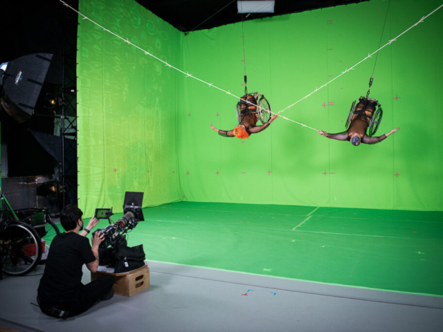 Alice Sheppard and Laurel Lawson soar in their wheelchairs in front of a bright green screen, each tipping sideways with arms extended. Laurel’s shadow flies next to her, on the right side green screen. Alice is a multiracial Black woman with short bright orange curly hair and coffee-colored skin; Laurel is a white person with pale skin and cropped peacock blue hair. They wear shimmery copper bodysuits overlaid with black mesh which shimmers in crimson, sapphire, and amethyst. They wear delicate filigree headpieces of copper and brass with pearl and gold beading. Photo by Cherylynn Tsushima.