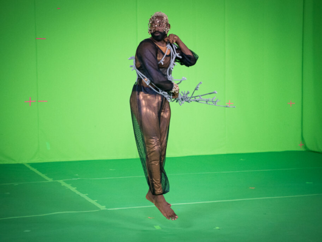 Jerron Herman jumps in the air, surrounded by a bright green screen and floor. His legs are together as he leaps, he looks over his shoulder as he whips a cloth wire prop at his waist and rests his other fist on the opposite shoulder. Jerron is a medium-build Black man with a dark beard and shortcut blonde hair. He wears a shimmery copper bodysuit overlaid with black mesh which shines in crimson, sapphire, and amethyst. He wears a delicate filigree headpiece of copper and brass with pearl and gold beading. Photo by Cherylynn Tsushima.