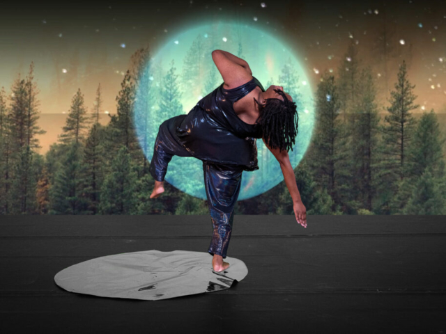 Kayla Hamilton stands on one leg in a mirrored pool; her back to the camera. Energy shoots into the ground as she arcs backward, lifts one leg and lets her head follow the pull of her opposite arm as it points to the ground. Behind her, a huge luminescent moon projection encircles her torso and a dense forest fills the background. Kayla is a Black woman with shoulder length locs; she wears a dark shimmery sleeveless tunic and pants. Photo Cherylynn Tsushima