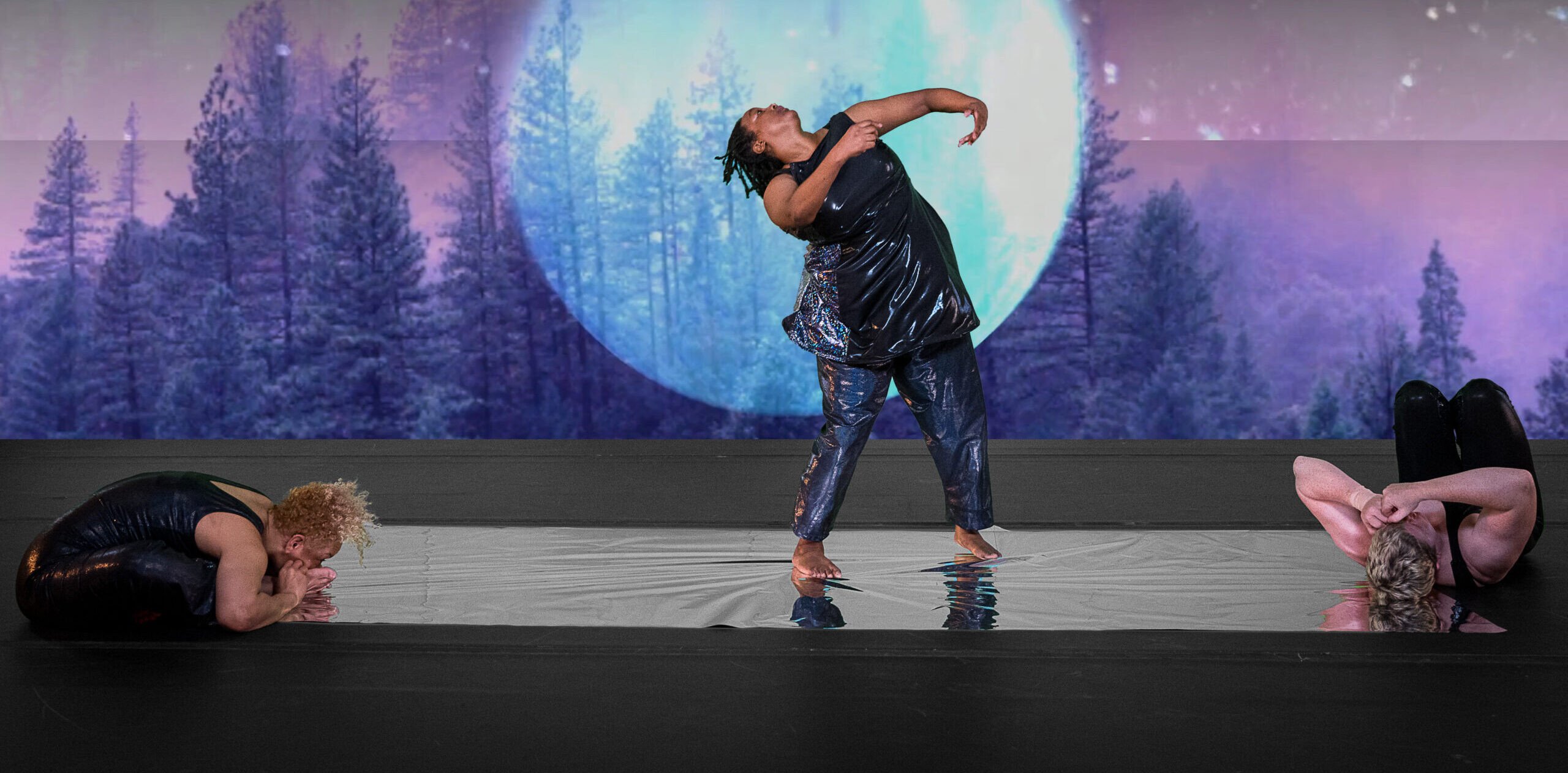 Kayla Hamilton stands on a reflective strip of silver, chin lifted as she arches her torso backwards, arms curved in front of her chest. Alice Sheppard sits on one end of the silver floor, torso folded into her own lap. Laurel Lawson lays on their back at the other end of the silver. Alice is a multiracial Black woman with short curly blonde hair; Kayla is a Black woman with shoulder length locs; Laurel is a white person with cropped hair. All three dancers wear dark shimmery bodysuits, a detailed video projection of a huge luminescent moon and a dense forest drenched in hues of cool blues and rich purples fills the background. Photo Cherylynn Tsushima