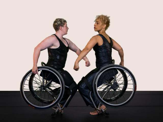 Laurel Lawson and Alice Sheppard are knee to knee, balancing on their footplates and lifting their wheels. They twist toward each other, arms curved, nearly hooking their left arms as they regard and mirror each other with intense expression. Laurel is a white person with cropped hair and pale freckled skin and Alice is a multiracial Black woman with coffee-colored skin and blonde curly hair piled atop her head; they both wear dark shimmery sleeveless bodysuits. The background is a soft cream tone.